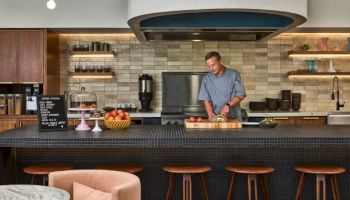A man works behind a modern bar with a curved hood, wooden stools, tiled backsplash, and a bowl of fruit on the counter.
