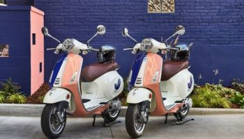 Two pastel-orange and cream scooters parked side by side on a driveway in front of a blue wall, with plants along the edge.