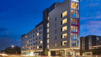 A modern multi-story hotel or apartment building with large glass windows, lit interiors, and a blue evening sky, along a busy street.