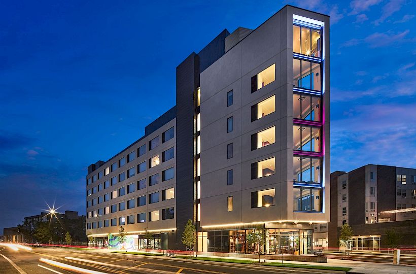 A modern multi-story hotel or apartment building with large glass windows, lit interiors, and a blue evening sky, along a busy street.
