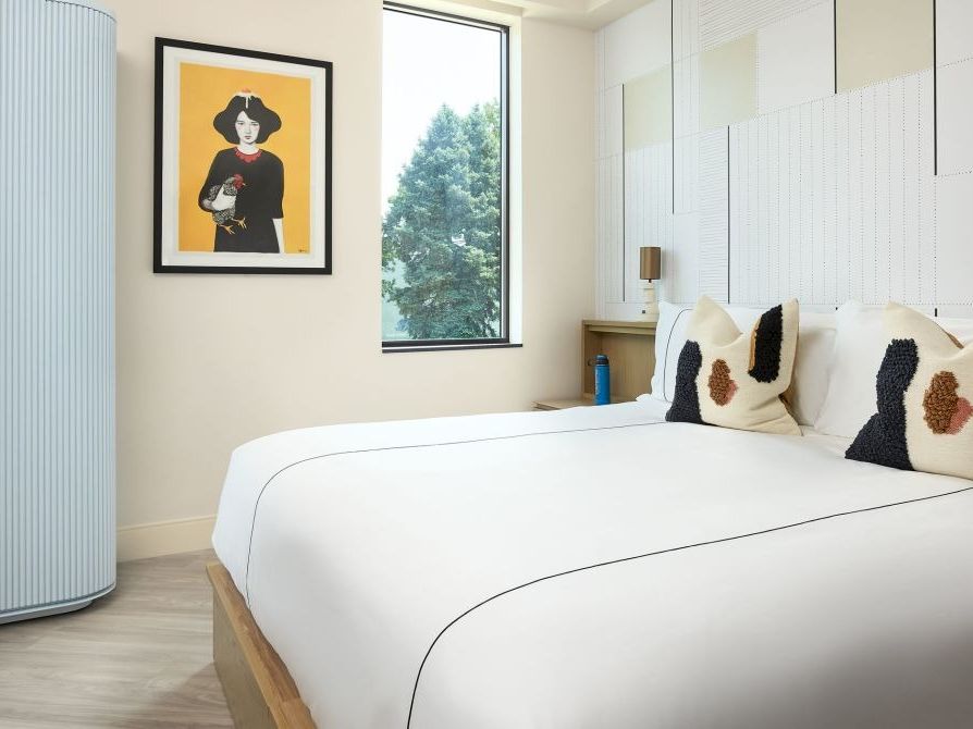 A modern hotel-style bedroom with a large white bed, decorative pillows, pale walls, a framed art piece, and a large window beside a curved cabinet, ending.