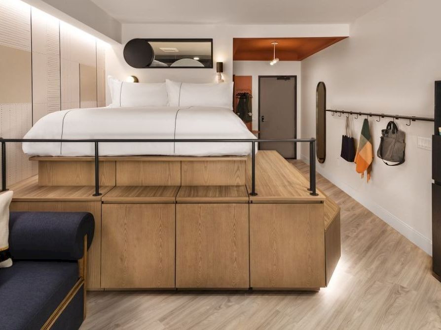 A compact, modern studio with a loft bed over wooden storage, a small sofa, wall hooks, and a minimalist kitchen area in warm wood tones.