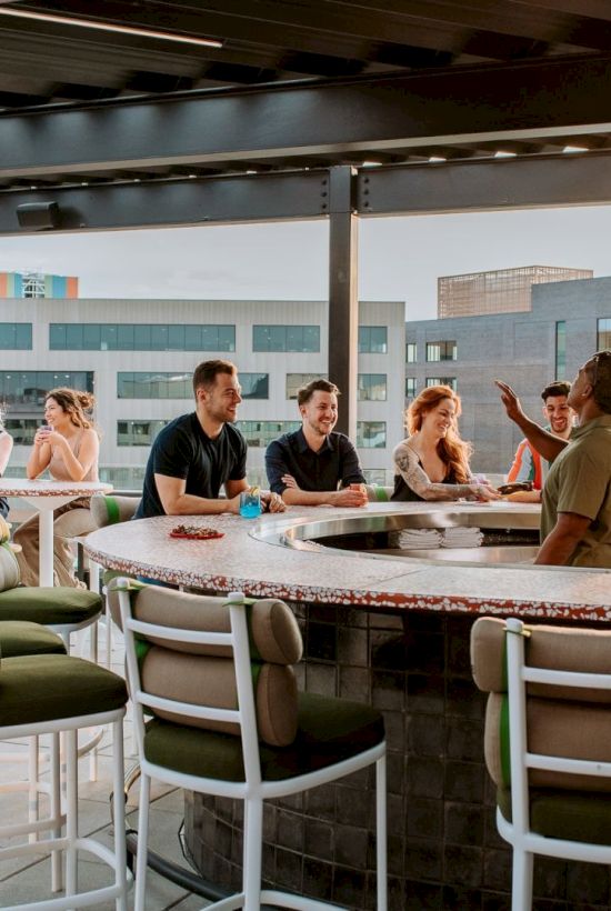 A group of people socializing around a high outdoor bar with stools, chatting and enjoying the rooftop setting.