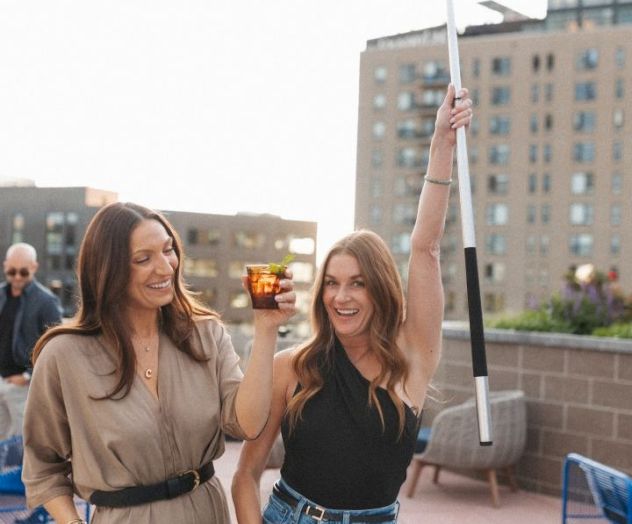 Two friends celebrate on a rooftop bar: one raises a bat and a drink, smiling, as they toast with a cityscape behind them.