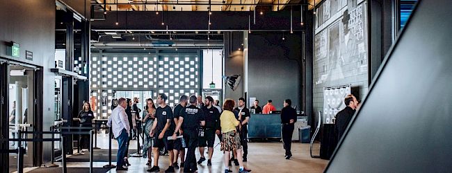 A modern indoor event with people mingling near a patterned wall, exposed ceiling beams, and warm lighting, as a casual networking scene.