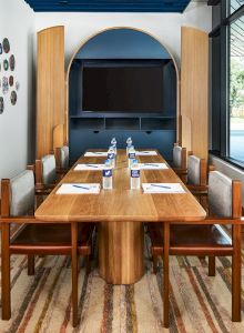 A modern dining room setup with a wooden table for eight, water glasses and place settings, art on the wall, and a large window view, bright and inviting. (140 chars)