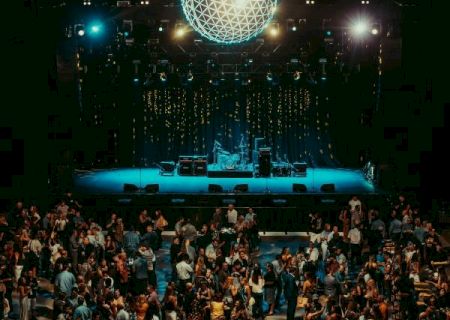 A crowded concert venue with a stage lit in blue, a geometric globe chandelier above, and people mingling under hanging lights.